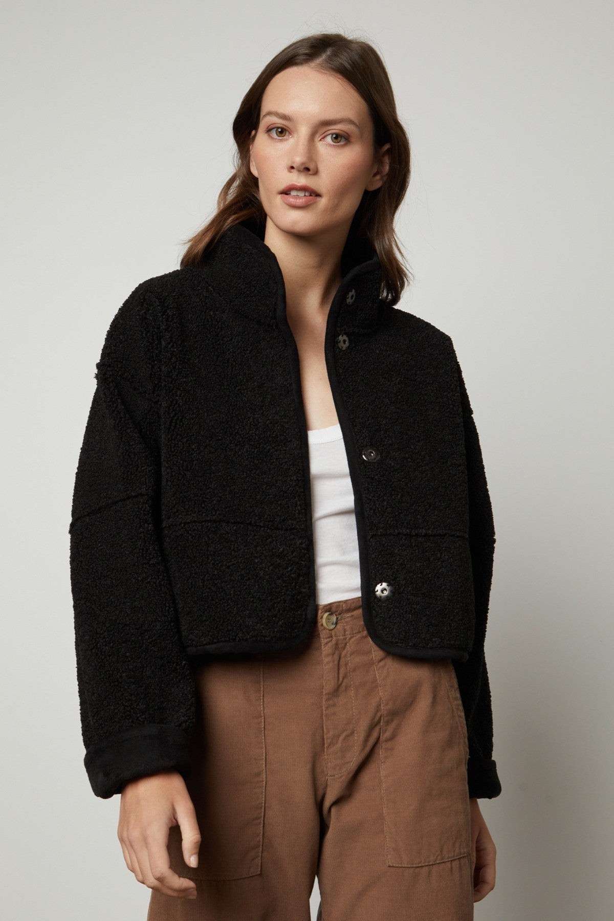 A woman stands against a plain light background wearing the Velvet by Graham & Spencer KELLY LUXE SHERPA REVERSIBLE JACKET in black over a white top and brown high-waisted pants.-40513965097153