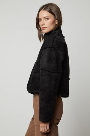 A woman stands sideways against a plain background, wearing Velvet by Graham & Spencer’s KELLY LUXE SHERPA REVERSIBLE JACKET in black with faux shearling details and brown pants.