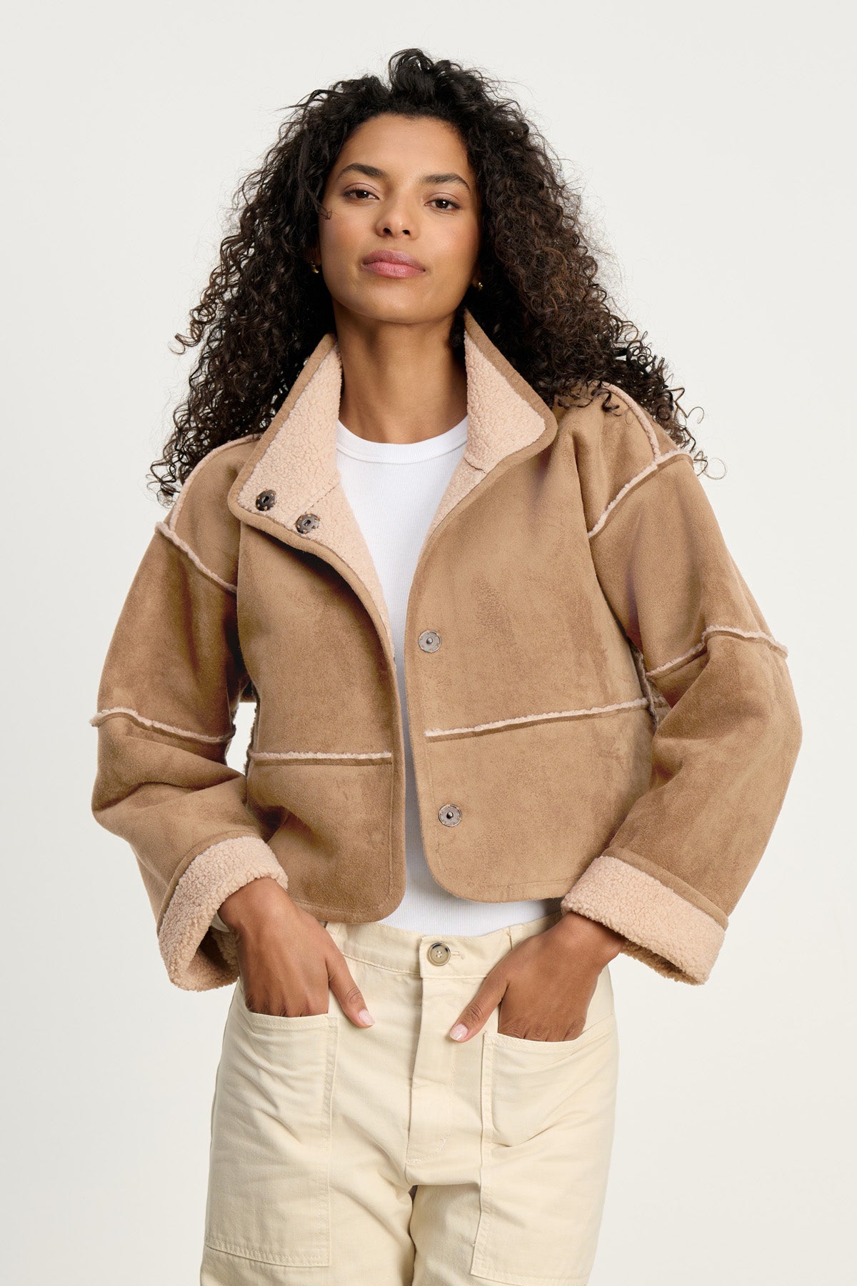 A woman stands with hands in pockets against a neutral background, wearing the Velvet by Graham & Spencer KELLY LUXE SHERPA REVERSIBLE JACKET in tan faux suede shearling, layered over a white shirt with light-colored pants.
