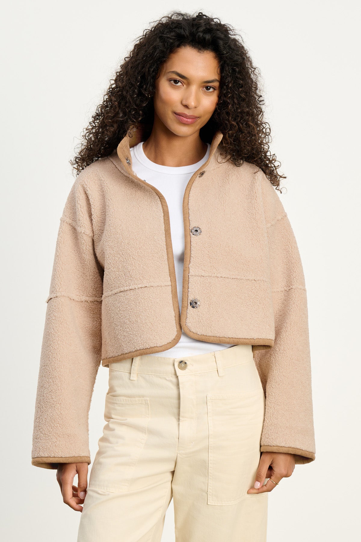 A woman with curly hair models the KELLY LUXE SHERPA REVERSIBLE JACKET by Velvet by Graham & Spencer over a white shirt and high-waisted beige pants, standing against a plain background.