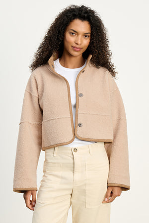 A woman with curly hair models the KELLY LUXE SHERPA REVERSIBLE JACKET by Velvet by Graham & Spencer over a white shirt and high-waisted beige pants, standing against a plain background.