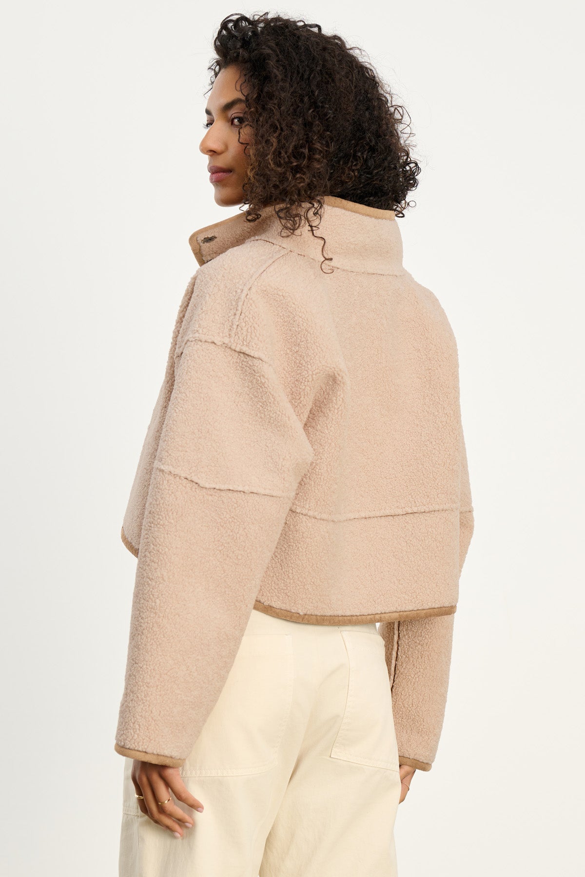 Model wears the Velvet by Graham & Spencer KELLY LUXE SHERPA REVERSIBLE JACKET in beige, featuring a cropped fit and high collar, standing sideways and looking over their shoulder against a plain background.