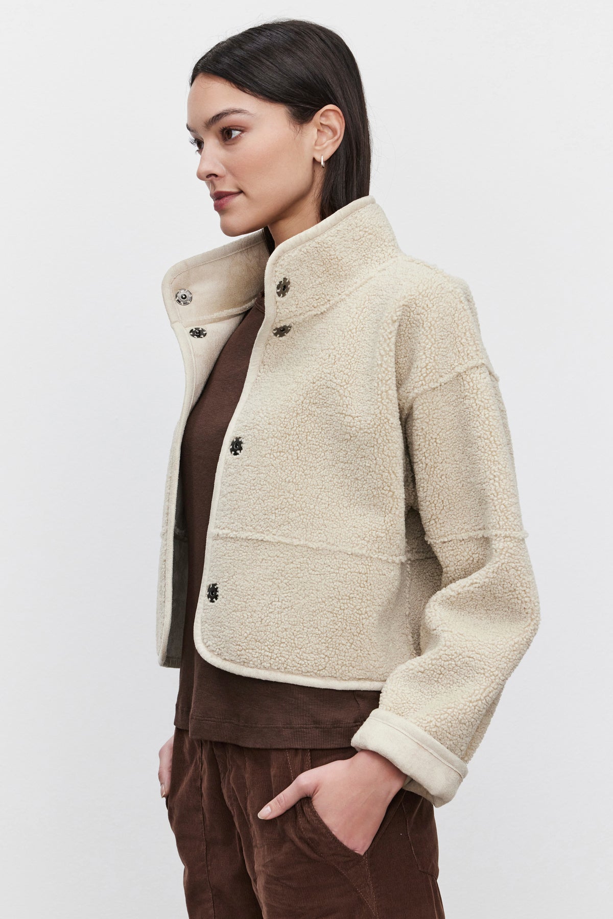   Side view of a person wearing the KELLY LUXE SHERPA REVERSIBLE JACKET by Velvet by Graham & Spencer, showcasing its elegant transition from a beige fleece to a stylish faux suede look, draped over a brown top with one hand casually in their pocket. 