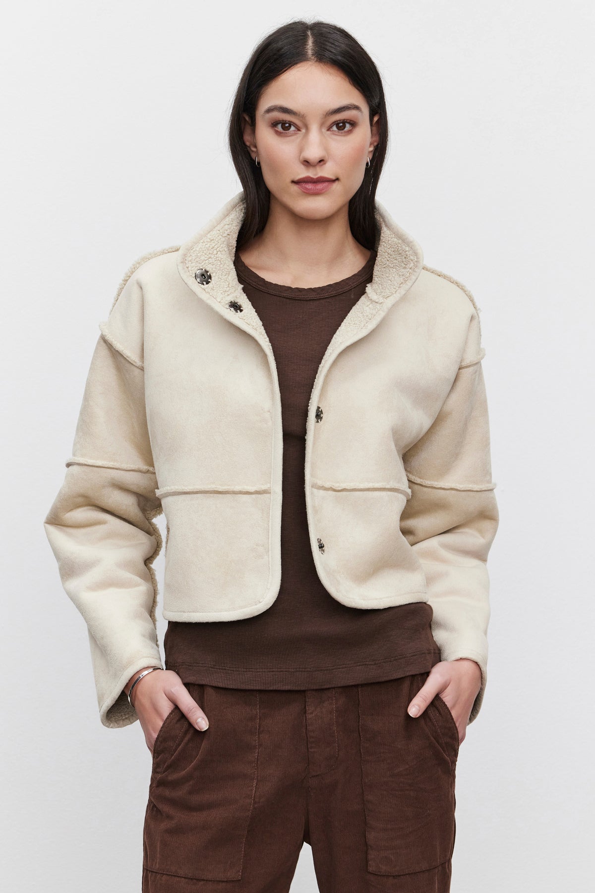   An individual is wearing the KELLY LUXE SHERPA REVERSIBLE JACKET by Velvet by Graham & Spencer, featuring beige faux suede and touches of faux sherpa, paired with a brown top and pants, against a plain background. 