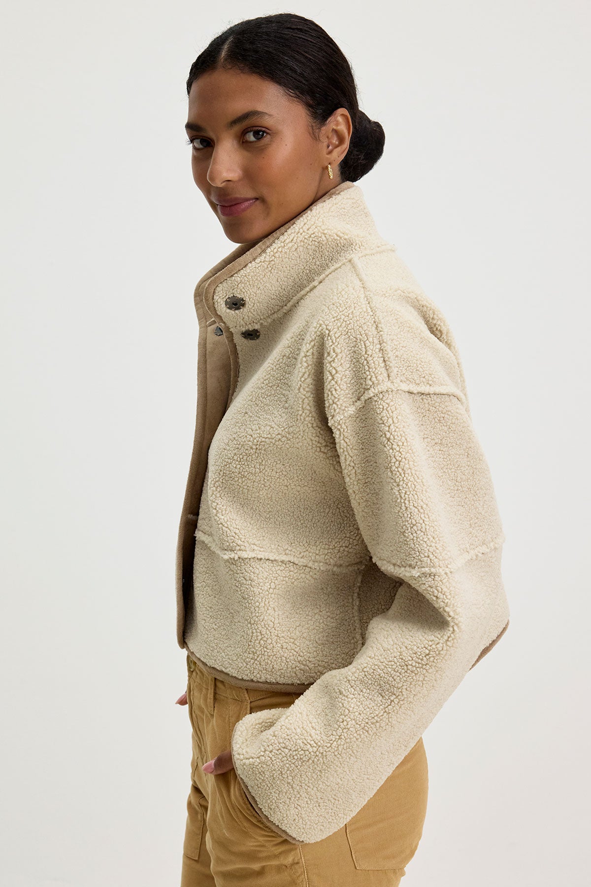   Wearing the KELLY LUXE SHERPA REVERSIBLE JACKET by Velvet by Graham & Spencer, an individual turns slightly to one side, highlighting its light beige fleece with a high collar and reversible faux sherpa design. 