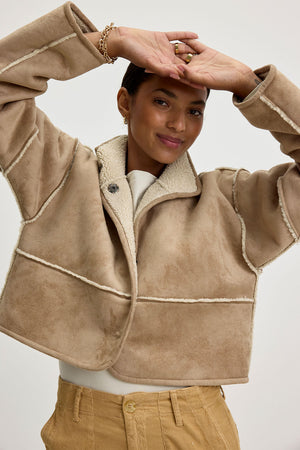 A person wearing the KELLY LUXE SHERPA REVERSIBLE JACKET by Velvet by Graham & Spencer, accompanied with tan pants, stands with arms raised against a plain background.