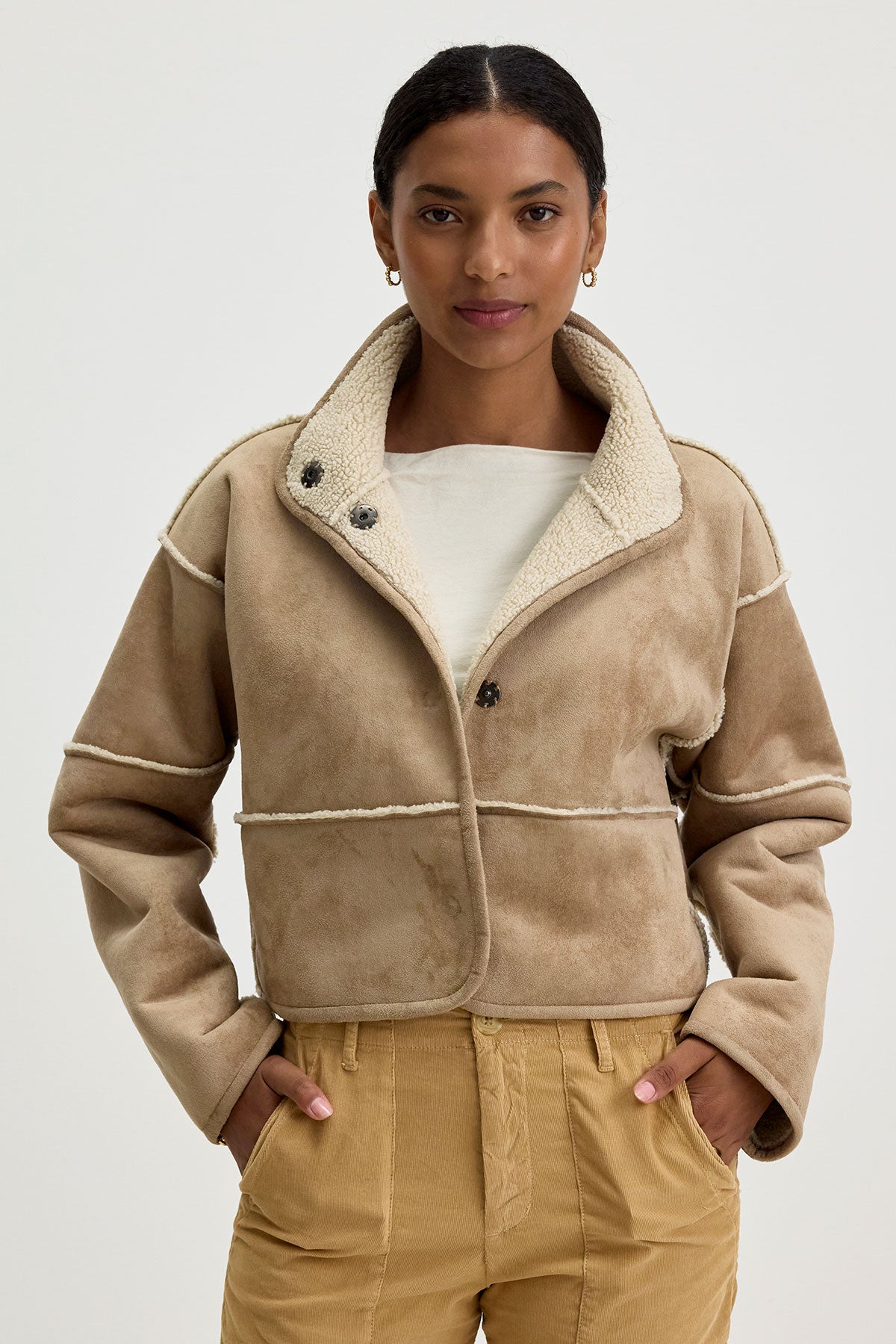   A person wearing the KELLY LUXE SHERPA REVERSIBLE JACKET by Velvet by Graham & Spencer, featuring a folded collar, paired with tan pants, standing with hands in pockets against a plain background. 