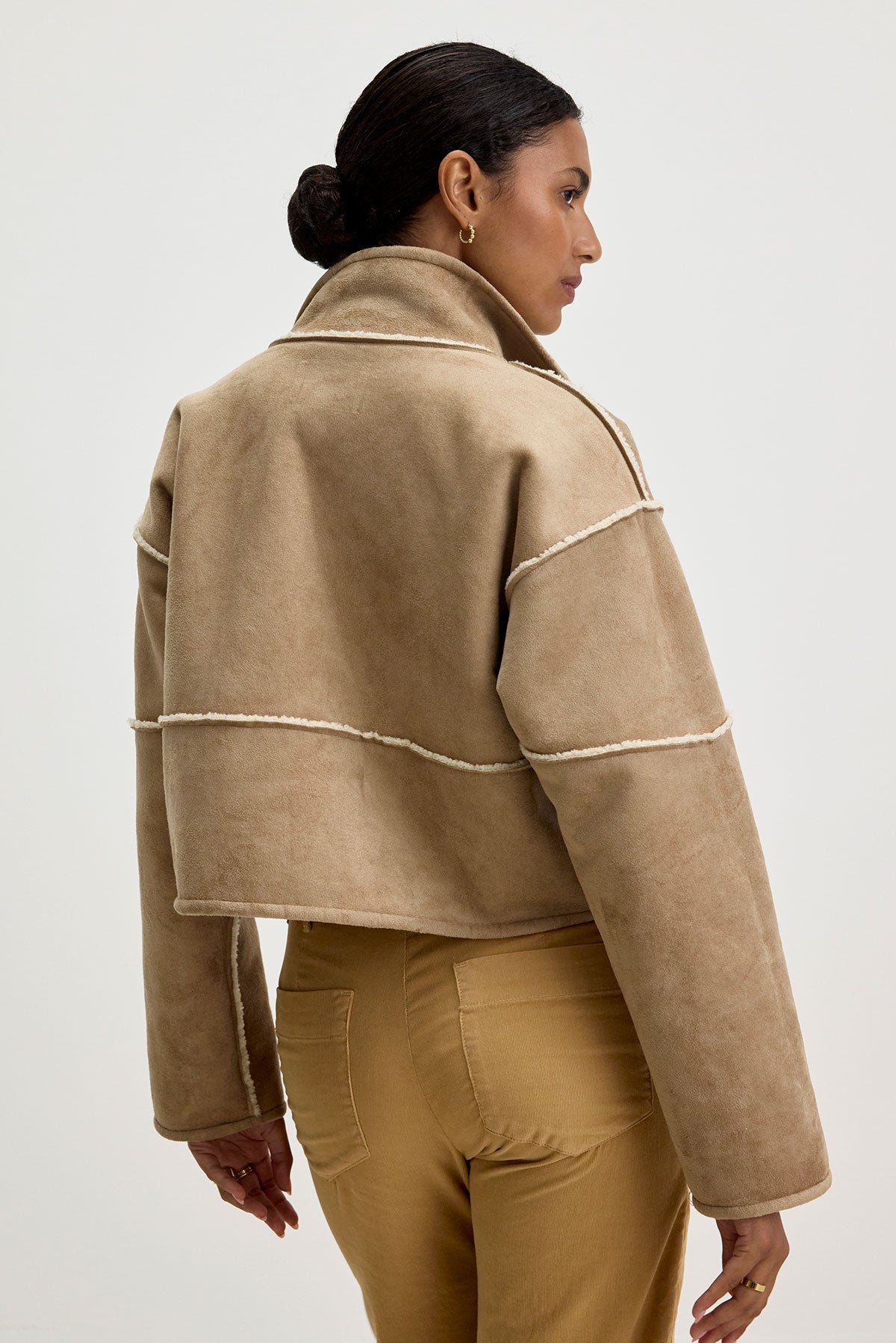   A person wearing the Velvet by Graham & Spencer KELLY LUXE SHERPA REVERSIBLE JACKET, which features a high collar and visible seam lines, is paired with tan pants and stands with their back to the camera. 