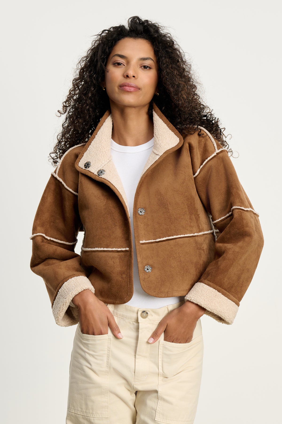 A woman stands with hands in pockets, wearing the KELLY LUXE SHERPA REVERSIBLE JACKET by Velvet by Graham & Spencer over a white shirt and cream pants against a plain light background.-39838356340929