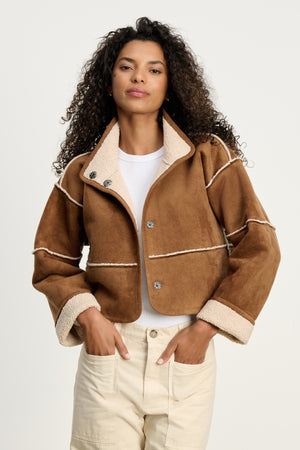 A woman stands with hands in pockets, wearing the KELLY LUXE SHERPA REVERSIBLE JACKET by Velvet by Graham & Spencer over a white shirt and cream pants against a plain light background.
