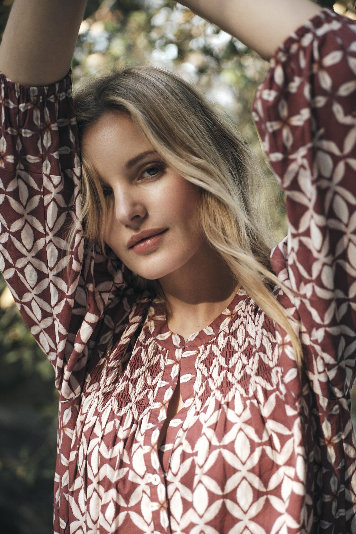   A person with long blonde hair wears the HADDON TOP by Velvet by Graham & Spencer, a patterned, long-sleeve shirt made of printed cotton cambric, and poses outdoors with their arms raised, looking at the camera. 