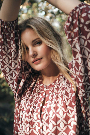 A person with long blonde hair wears the HADDON TOP by Velvet by Graham & Spencer, a patterned, long-sleeve shirt made of printed cotton cambric, and poses outdoors with their arms raised, looking at the camera.