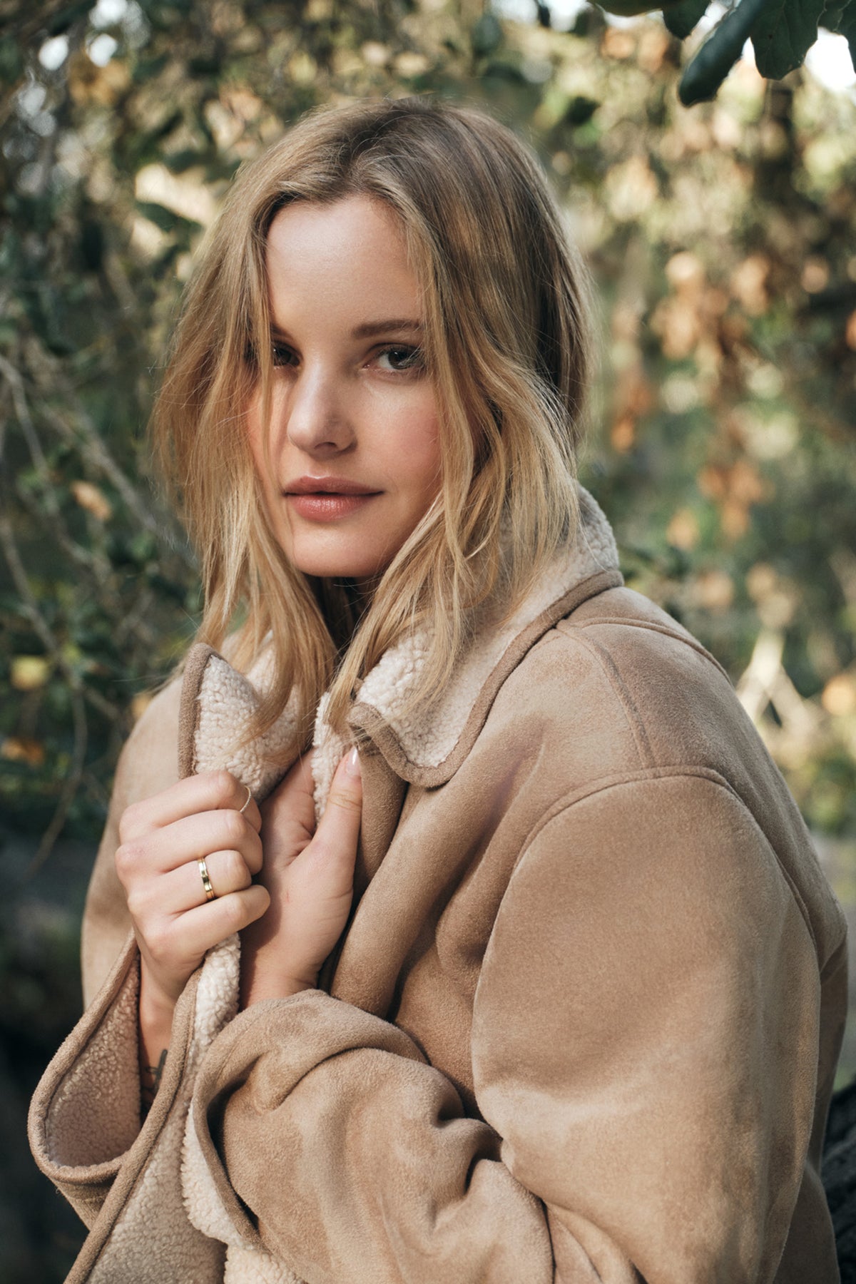 A person with long blonde hair stands outdoors, clutching the collar of a MOLLY LUXE SHERPA JACKET by Velvet by Graham & Spencer, surrounded by trees in the background.-37894005653697