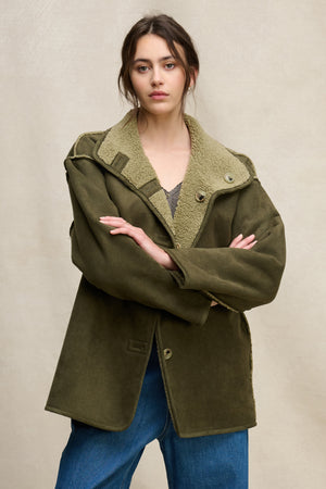 A woman stands with arms crossed, wearing the Velvet by Graham & Spencer SHONA SHERPA REVERSIBLE JACKET in oversized olive green over a gray top and blue jeans, set against a plain beige background.