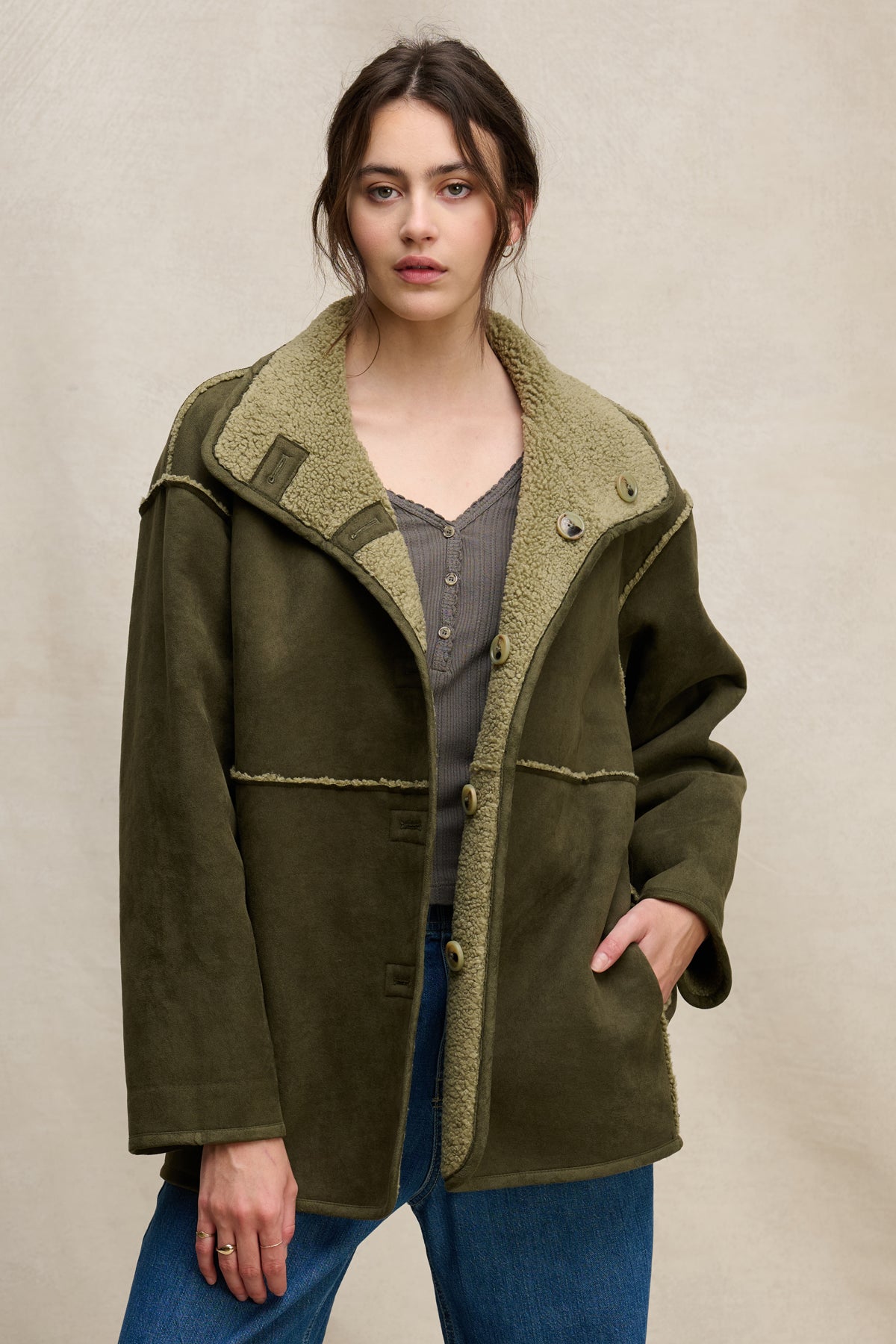 A woman wears the SHONA SHERPA REVERSIBLE JACKET by Velvet by Graham & Spencer in olive green over a gray top and blue jeans, standing against a neutral background.-39800405786817