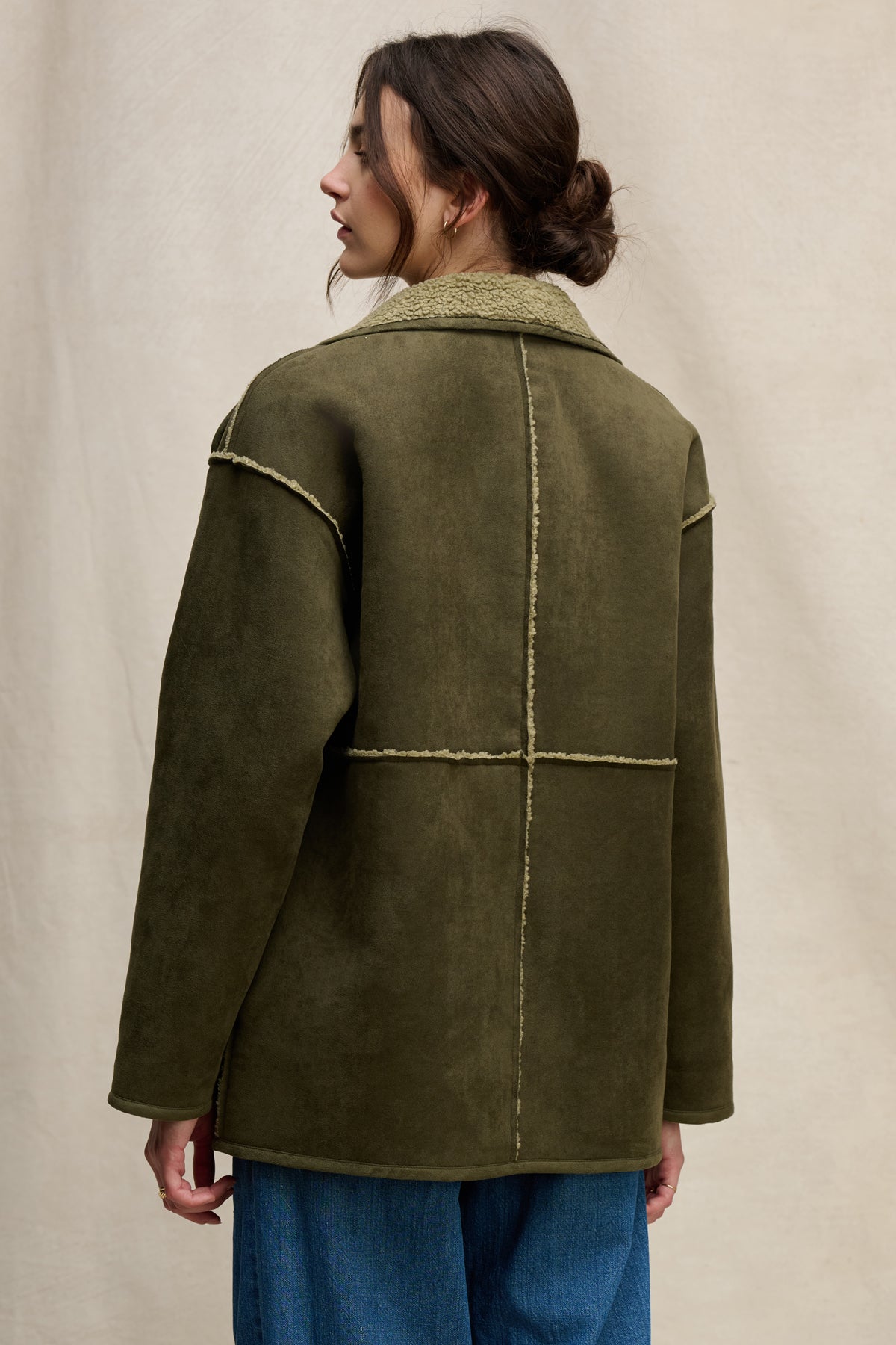 A person wears the SHONA SHERPA REVERSIBLE JACKET by Velvet by Graham & Spencer in olive green with blue jeans, standing facing away against a neutral background.-39800405688513