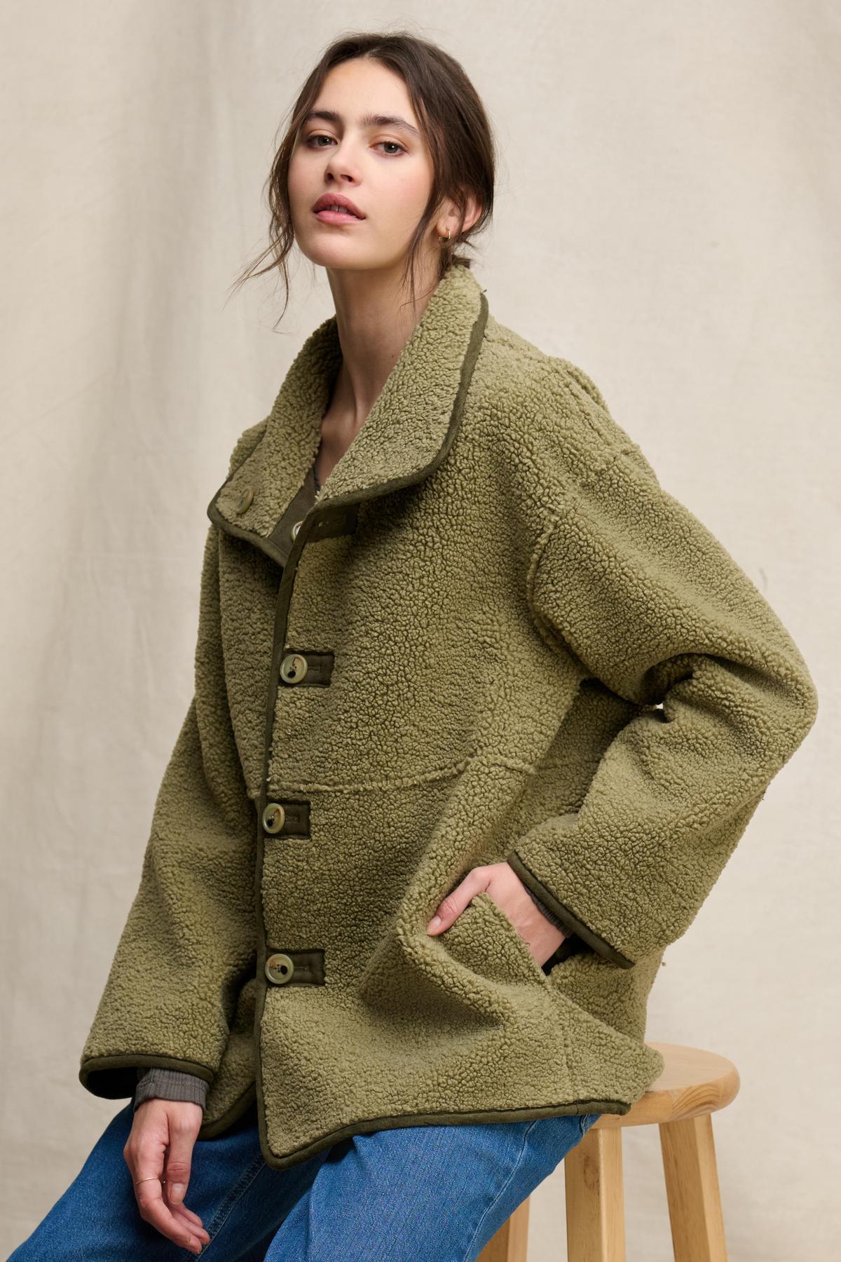   A person wearing the SHONA SHERPA REVERSIBLE JACKET by Velvet by Graham & Spencer, an oversized olive green faux sherpa fleece coat with large buttons, sits on a wooden stool against a neutral background. 