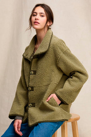 A person wearing the SHONA SHERPA REVERSIBLE JACKET by Velvet by Graham & Spencer, an oversized olive green faux sherpa fleece coat with large buttons, sits on a wooden stool against a neutral background.