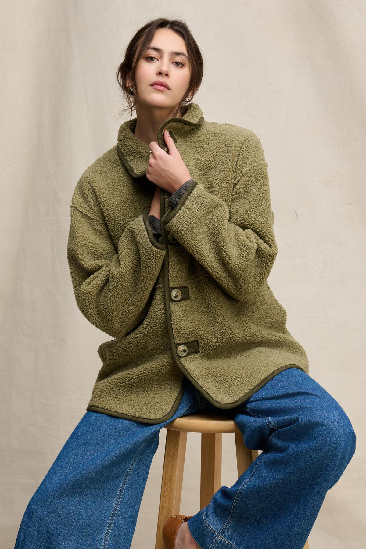   A person wearing the SHONA SHERPA REVERSIBLE JACKET by Velvet by Graham & Spencer in olive green and wide-leg blue jeans sits on a wooden stool against a neutral fabric backdrop. 