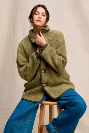 A person wearing the SHONA SHERPA REVERSIBLE JACKET by Velvet by Graham & Spencer in olive green and wide-leg blue jeans sits on a wooden stool against a neutral fabric backdrop.