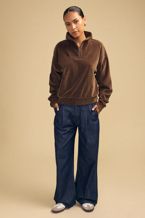 A person wearing a brown DANICA VELOUR HALF-ZIP SWEATSHIRT and wide-leg dark denim jeans.