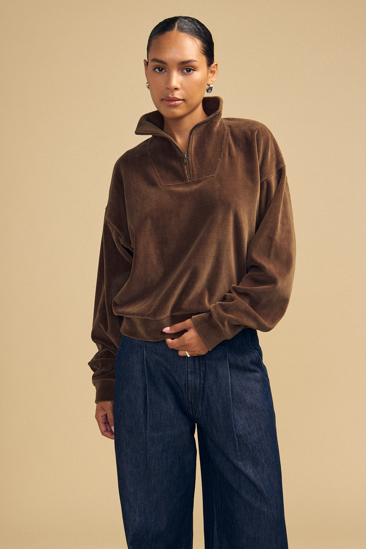 A person wears a brown DANICA VELOUR HALF-ZIP SWEATSHIRT and dark denim trousers.-46488235671745