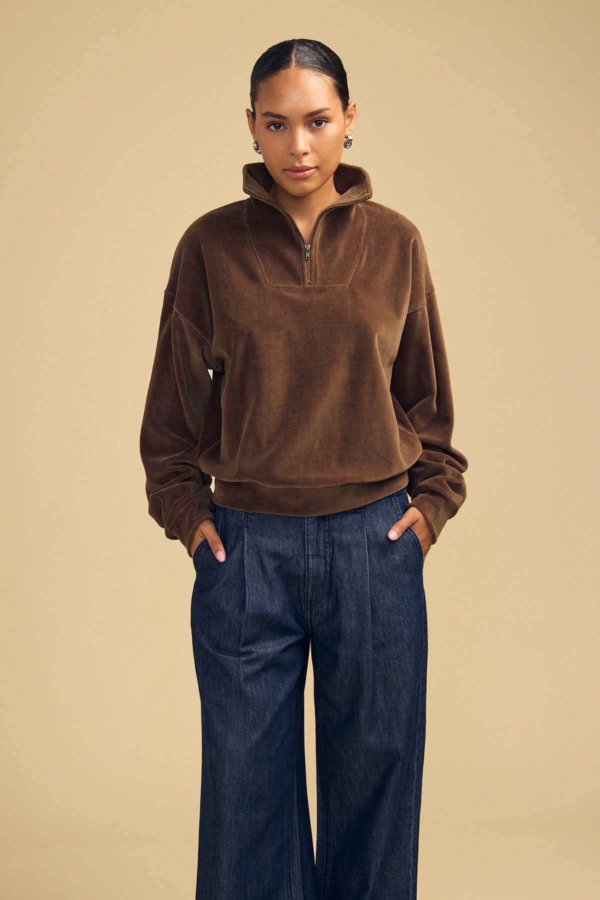 A person wearing a brown DANICA VELOUR HALF-ZIP SWEATSHIRT and dark blue denim trousers.-46488235704513