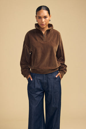 A person wearing a brown DANICA VELOUR HALF-ZIP SWEATSHIRT and dark blue denim trousers.