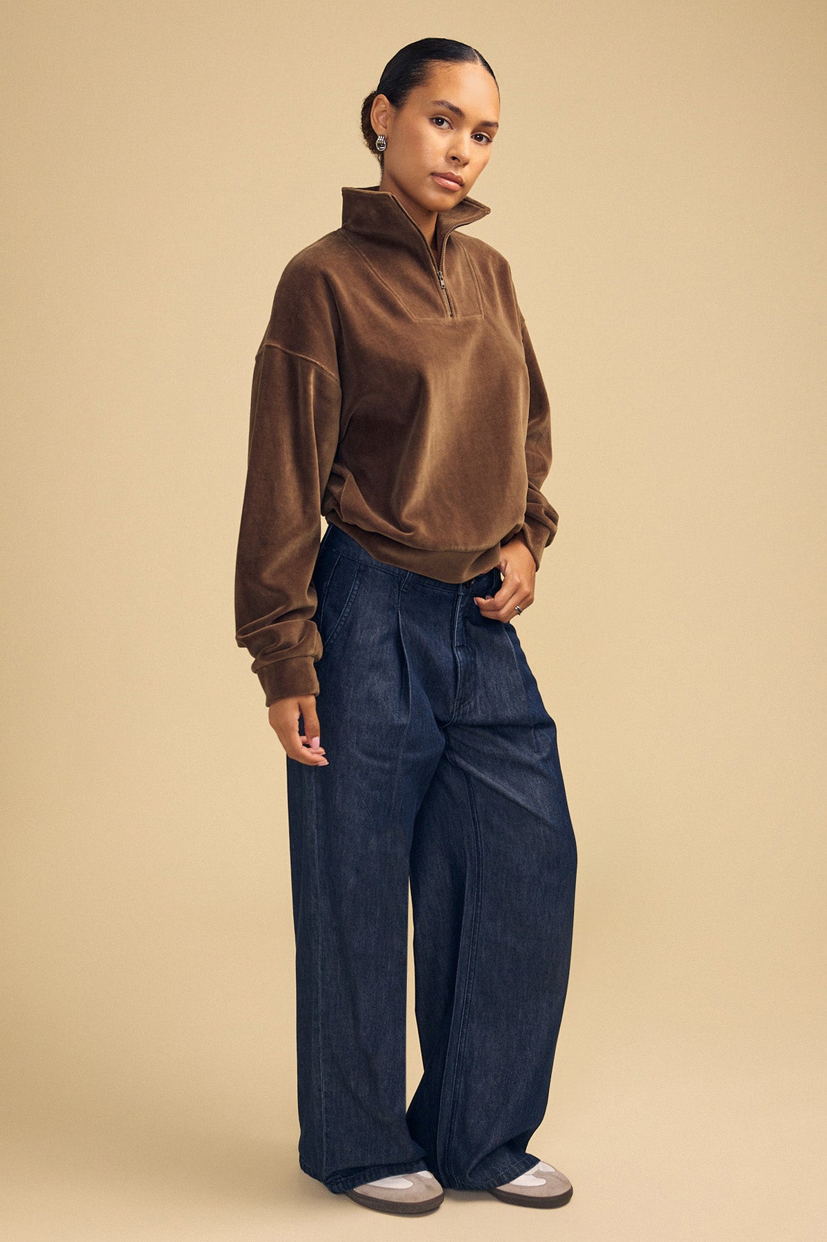 A person wearing a brown DANICA VELOUR HALF-ZIP SWEATSHIRT and wide-leg dark denim jeans.