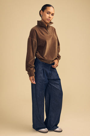 A person wearing a brown DANICA VELOUR HALF-ZIP SWEATSHIRT and wide-leg dark denim jeans.