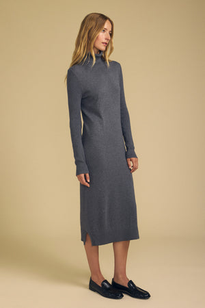 A woman stands against a plain beige background wearing the BEA COTTON CASHMERE DRESS by Velvet by Graham & Spencer—a long-sleeved, gray, midi-length sweater dress—paired with black loafers.