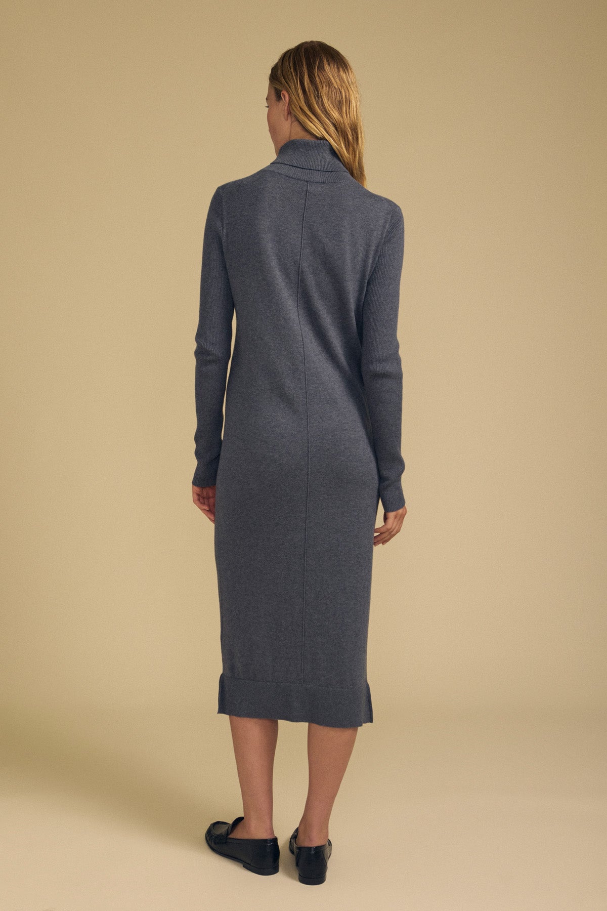 A person with long hair stands facing away, wearing the BEA COTTON CASHMERE DRESS by Velvet by Graham & Spencer—a long-sleeve, knee-length sweater dress—paired with black shoes against a plain beige background.-42063723069633