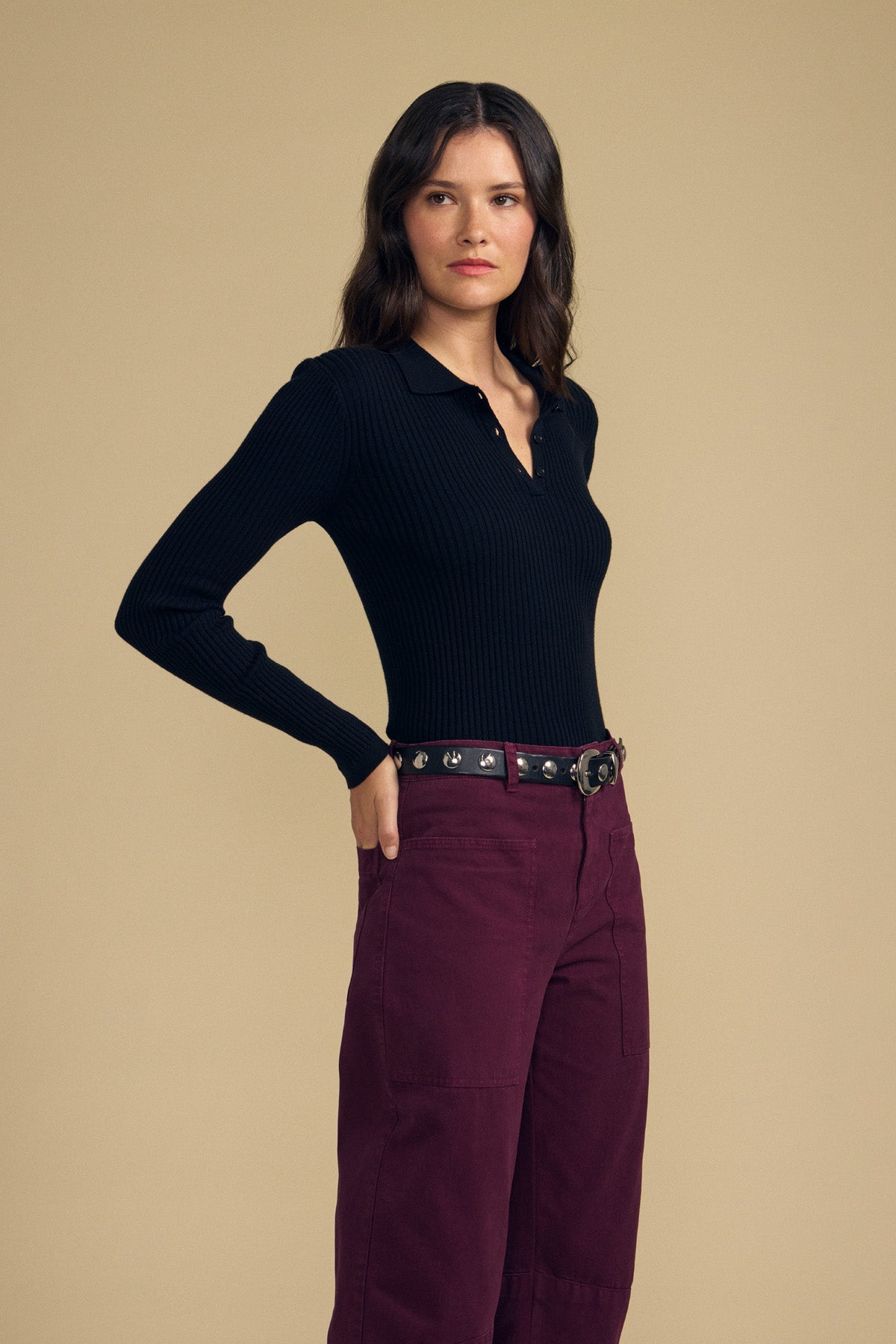 A woman with wavy brown hair stands against a plain beige background, wearing the FRANCES SWEATER POLO by Velvet by Graham & Spencer—a black, long-sleeve ribbed top—paired with maroon pants.-41815093772481