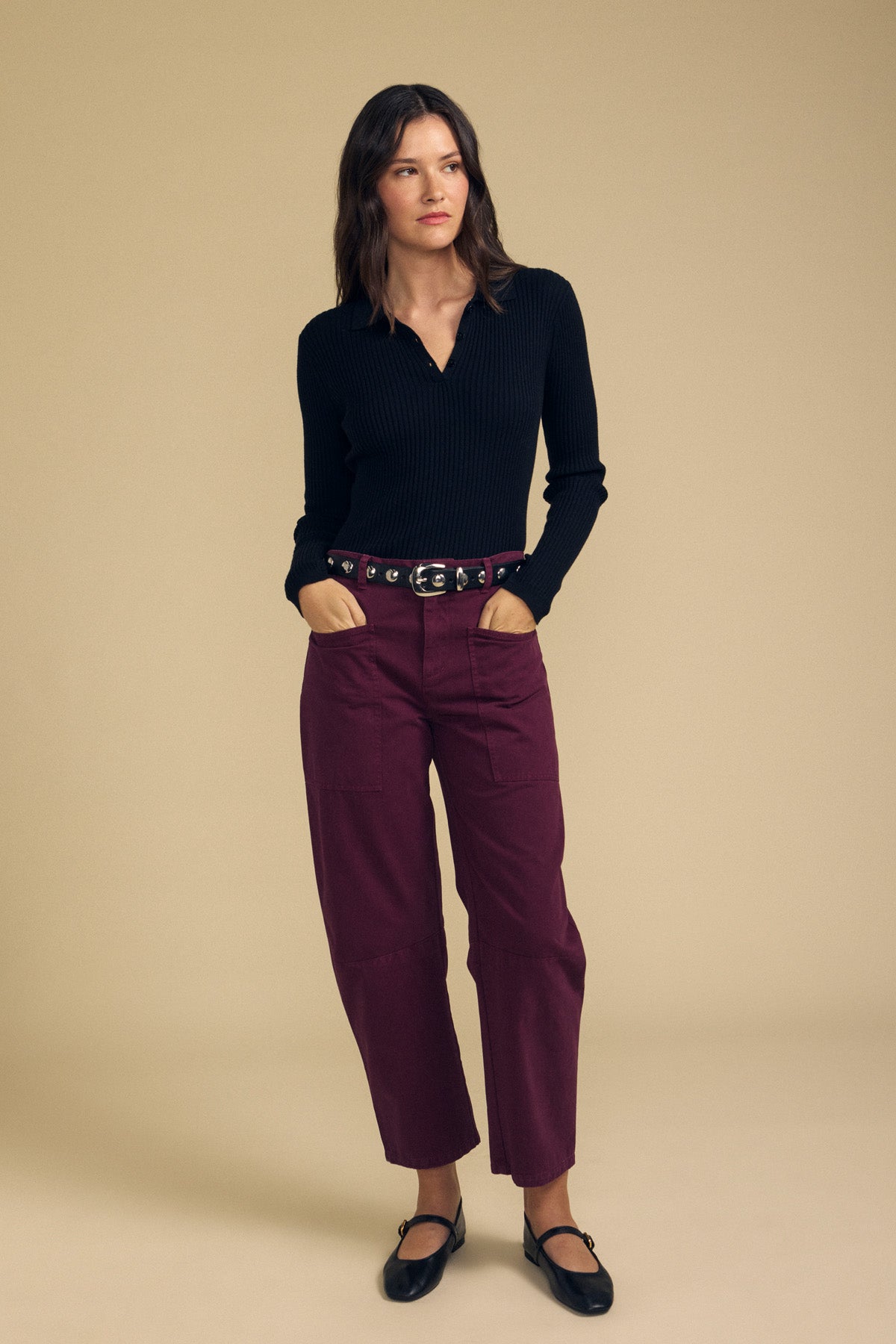 A woman stands against a beige background, wearing the FRANCES SWEATER POLO by Velvet by Graham & Spencer—a black long-sleeve top—paired with maroon pants, a black belt, and black strappy shoes.
