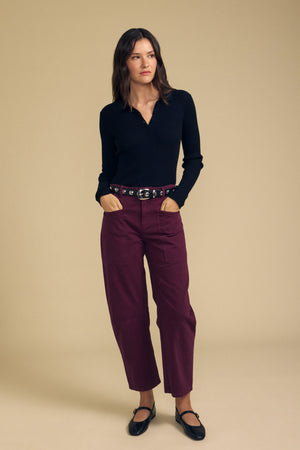 A woman stands against a beige background, wearing the FRANCES SWEATER POLO by Velvet by Graham & Spencer—a black long-sleeve top—paired with maroon pants, a black belt, and black strappy shoes.