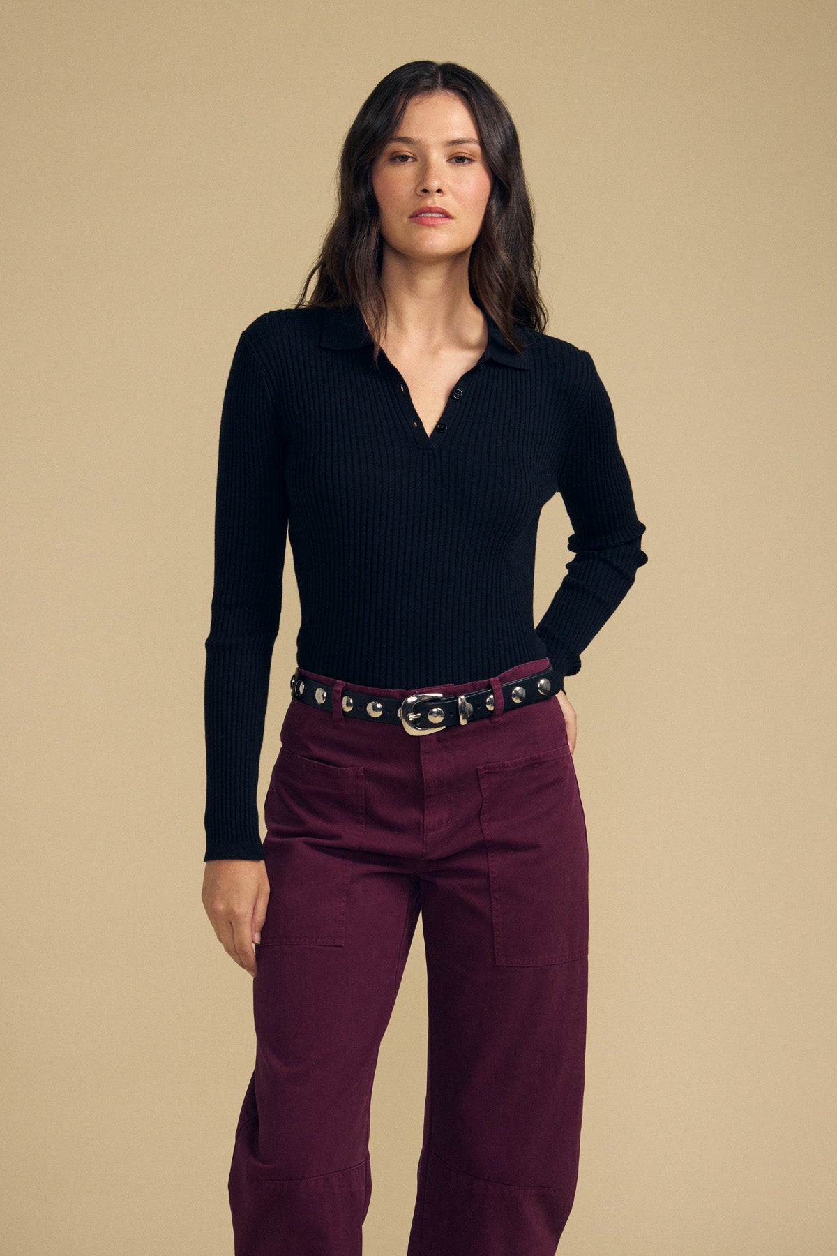 A woman stands against a beige background wearing the FRANCES SWEATER POLO by Velvet by Graham & Spencer, a black long-sleeve ribbed top, paired with high-waisted burgundy pants and a black belt.