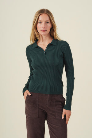 A woman stands against an off-white background, wearing the FRANCES SWEATER POLO in dark green from Velvet by Graham & Spencer's premium women's knitwear collection, paired with brown pants. She looks at the camera with a neutral expression.