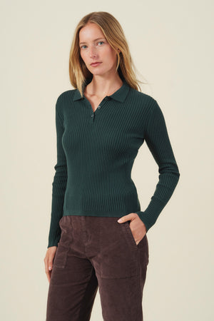 A woman stands facing forward, wearing the FRANCES SWEATER POLO by Velvet by Graham & Spencer—a chic dark green women's knitwear piece—paired with brown corduroy pants against a plain light background.