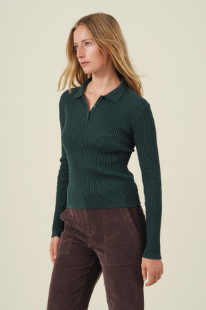A woman wears the FRANCES SWEATER POLO by Velvet by Graham & Spencer—a dark green, ribbed, long-sleeve sweater polo—paired with brown corduroy pants, standing against a plain light background. This stylish piece highlights the best in women’s knitwear.