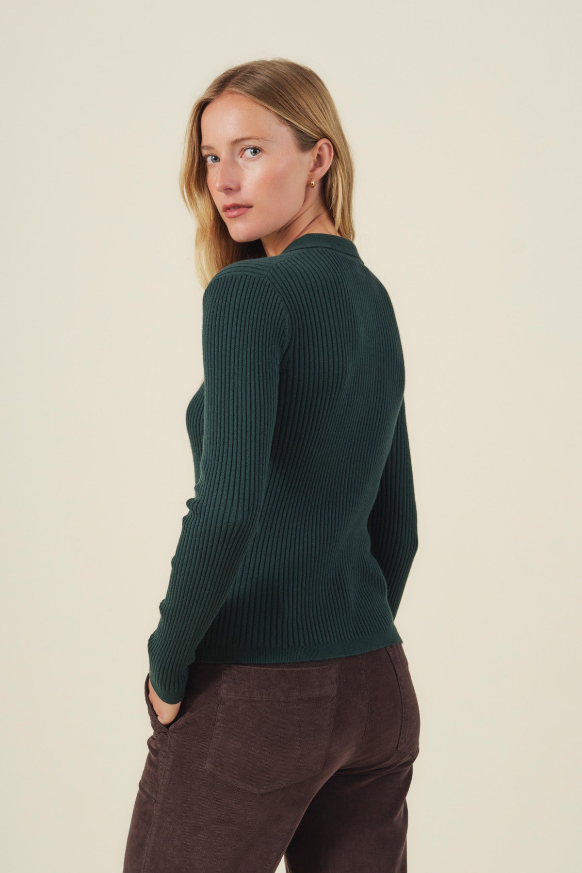 A woman with long blonde hair wears the Velvet by Graham & Spencer FRANCES SWEATER POLO in dark green, paired with brown pants, standing with her back turned and looking over her shoulder against a plain light background.