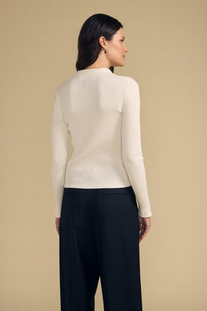 A woman stands with her back to the camera, wearing the FRANCES SWEATER POLO by Velvet by Graham & Spencer—a long-sleeve, ribbed white top—paired with black pants against a plain beige background.