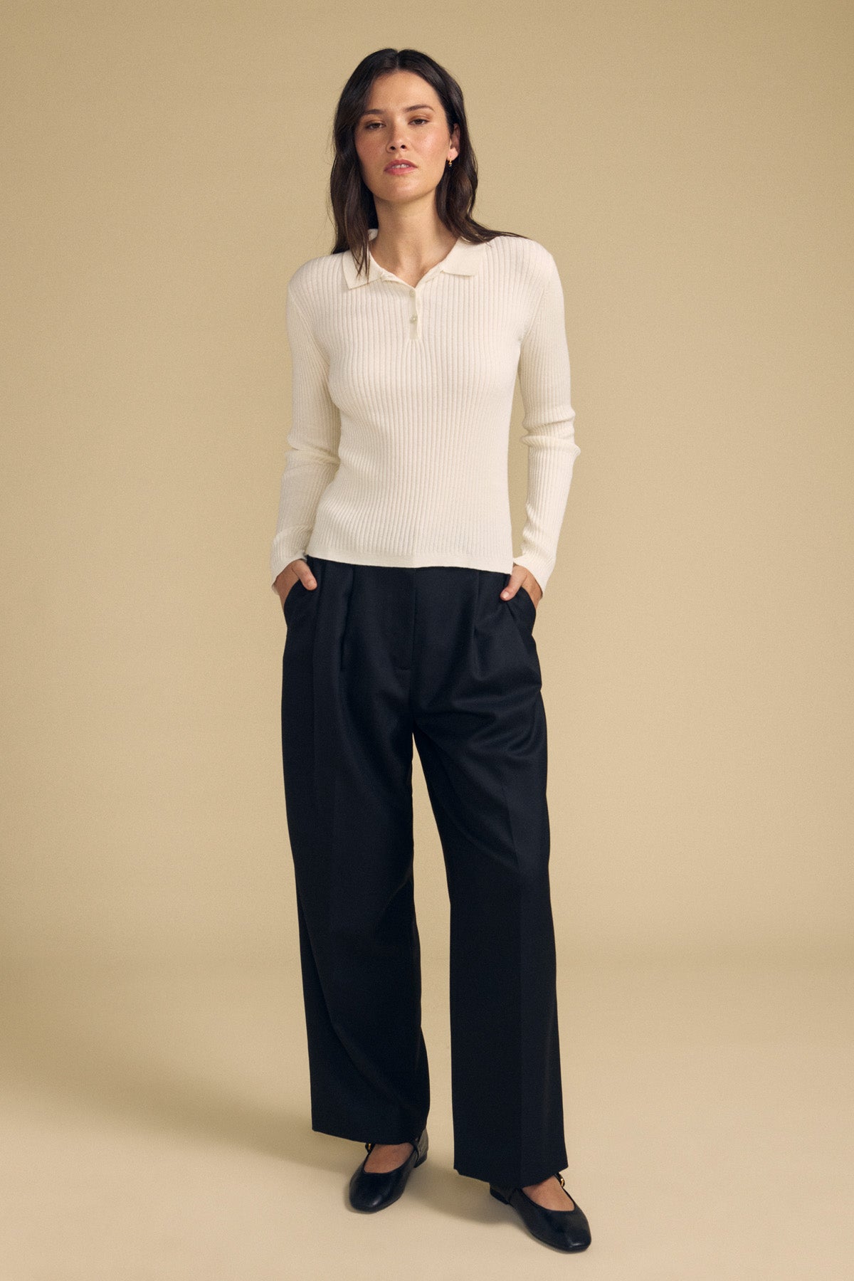 A woman stands against a beige background wearing the FRANCES SWEATER POLO by Velvet by Graham & Spencer—a cream ribbed long-sleeve top—paired with black wide-leg trousers and black flats, with her hands in her pockets.