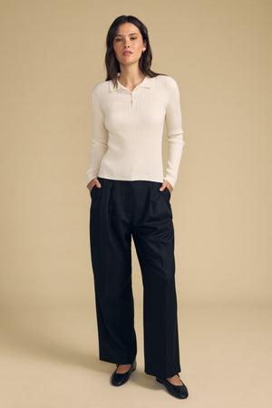 A woman stands against a beige background wearing the FRANCES SWEATER POLO by Velvet by Graham & Spencer—a cream ribbed long-sleeve top—paired with black wide-leg trousers and black flats, with her hands in her pockets.