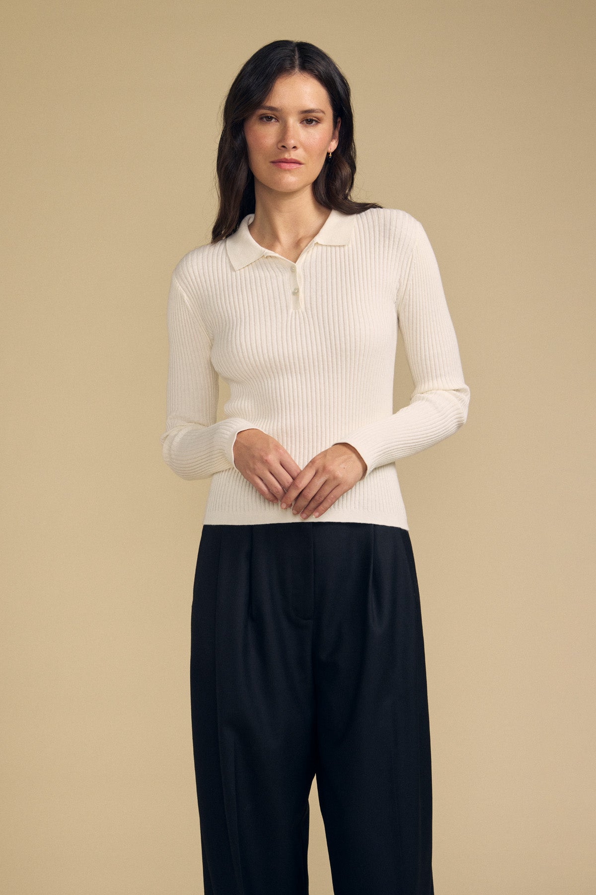 A woman stands against a beige background, wearing the FRANCES SWEATER POLO by Velvet by Graham & Spencer—a long-sleeve, cream ribbed polo sweater—paired with loose black trousers, hands resting in front.-41815093936321