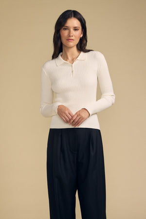 A woman stands against a beige background, wearing the FRANCES SWEATER POLO by Velvet by Graham & Spencer—a long-sleeve, cream ribbed polo sweater—paired with loose black trousers, hands resting in front.