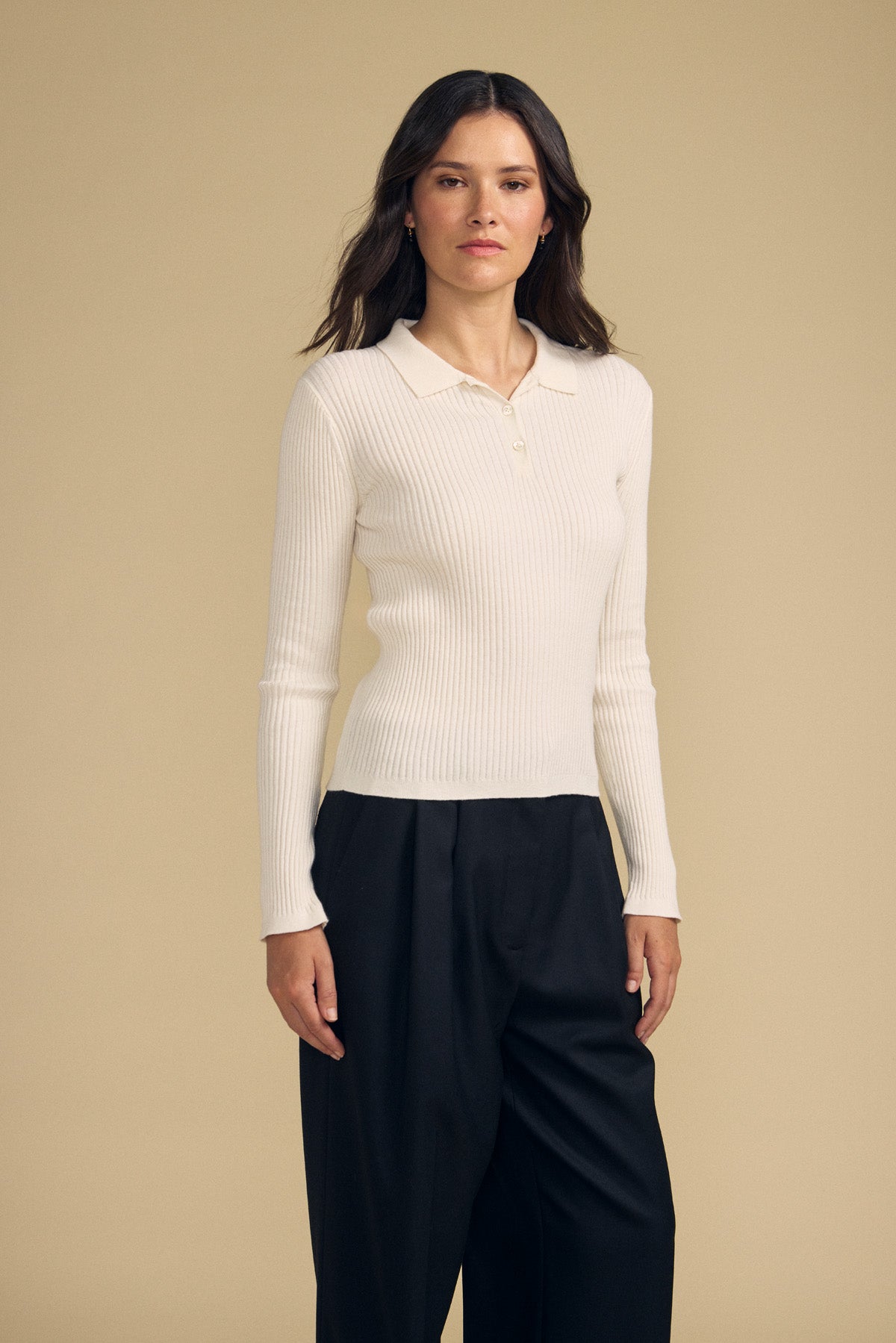 A woman stands against a beige background, wearing the FRANCES SWEATER POLO by Velvet by Graham & Spencer—a long-sleeved, ribbed cream polo shirt—paired with loose black trousers.-41815093903553