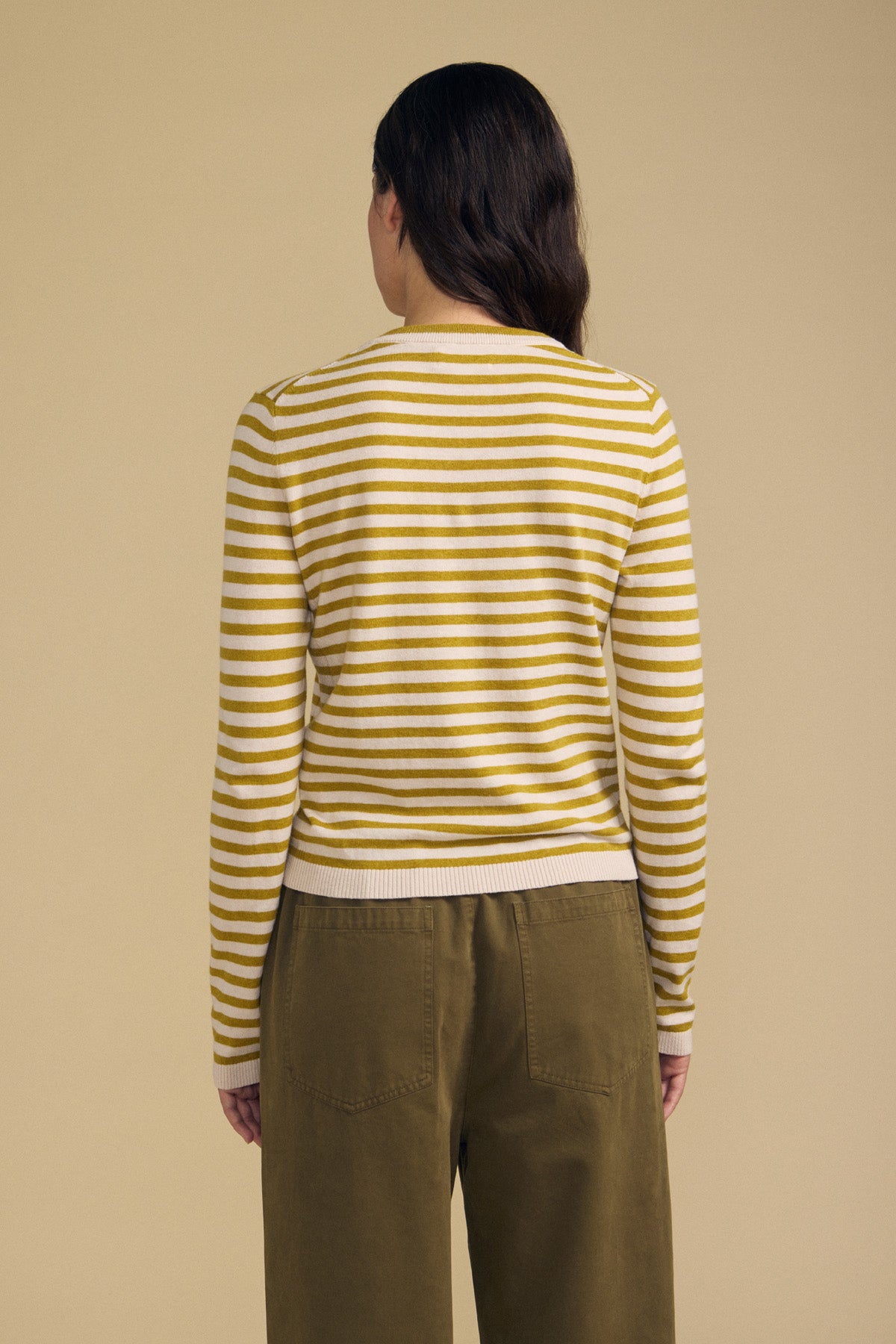 A person stands with their back to the camera, wearing the ROXY SWEATER by Velvet by Graham & Spencer in yellow and white stripes, paired with olive green pants against a plain beige background.