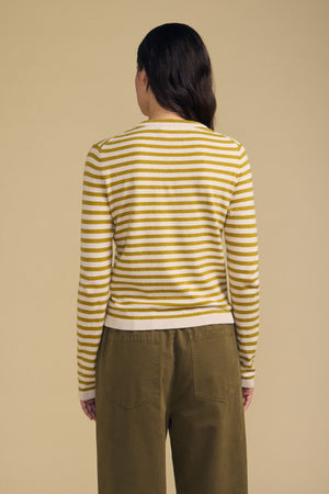 A person stands with their back to the camera, wearing the ROXY SWEATER by Velvet by Graham & Spencer in yellow and white stripes, paired with olive green pants against a plain beige background.
