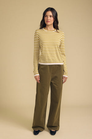 A woman stands against a beige background, wearing the ROXY SWEATER by Velvet by Graham & Spencer—featuring yellow and white stripes—paired with loose olive-green pants and black shoes.