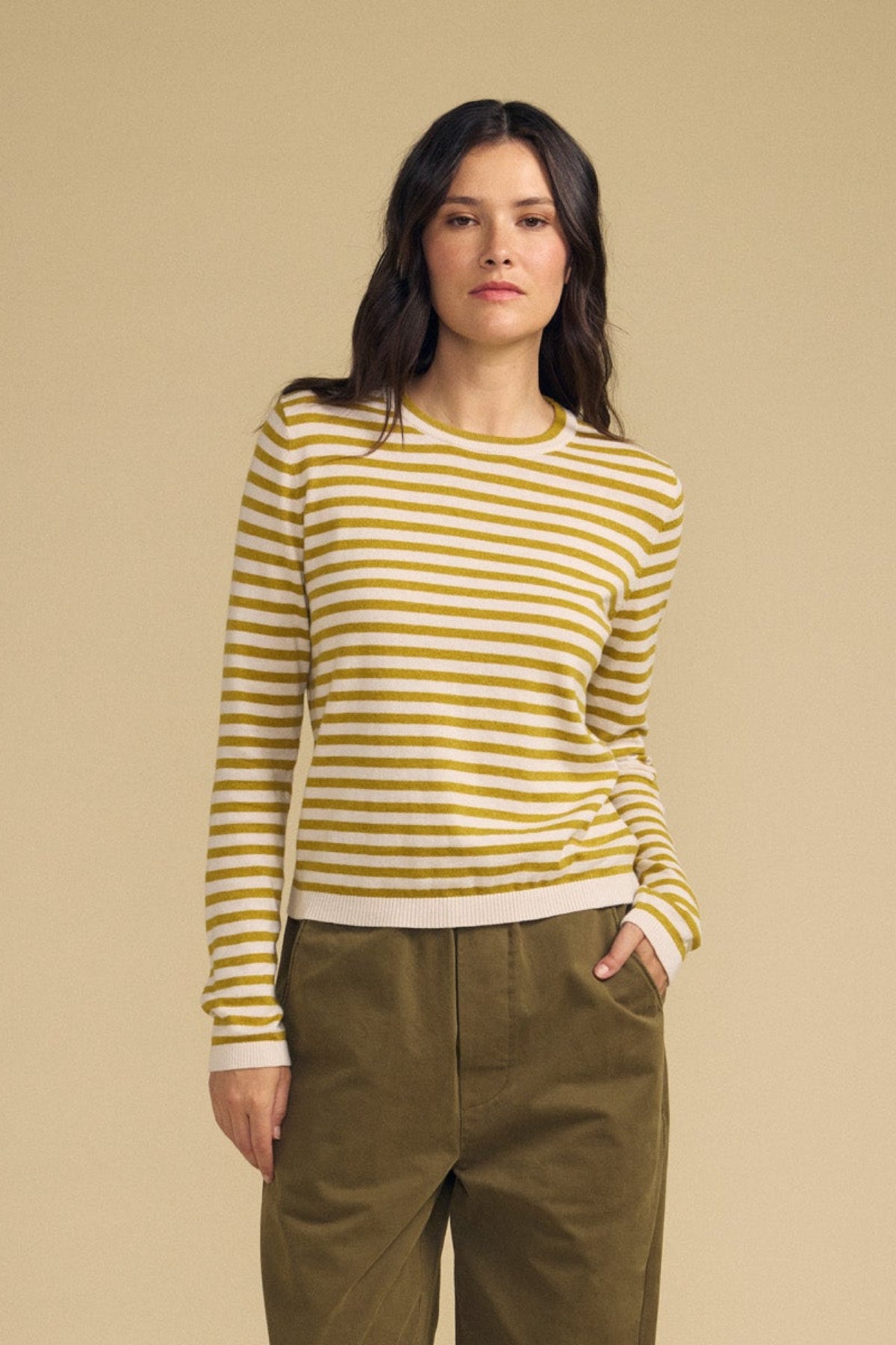 A person wearing the ROXY SWEATER by Velvet by Graham & Spencer, featuring yellow and white stripes, paired with olive green pants and standing against a plain beige background.